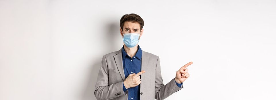 Covid-19, pandemic and business concept. Upset man in medical mask and office suit, frowning sad and pointing left at banner, standing on white background.