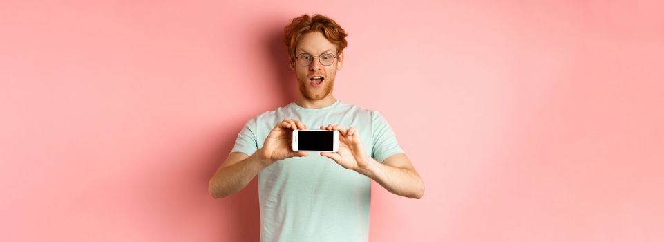 Amazed redhead man gasping and staring with awe phone, showing blank smartphone screen horizontally, standing over pink background.