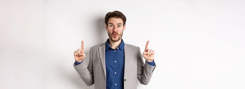 Excited businessman in suit say wow and smiling amused, pointing fingers up at good deal, standing against white background.