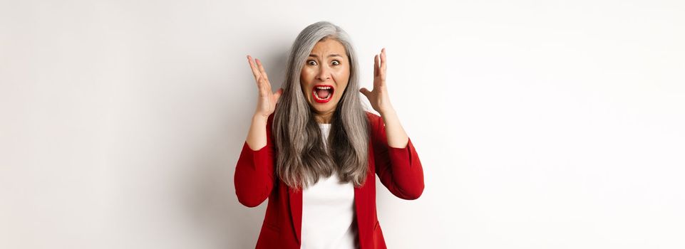 Asian senior businesswoman screaming and looking outraged, feeling distressed and shouting, standing against white background.