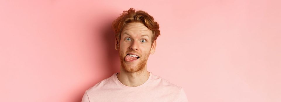 Headshot of funny redhead guy showing tongue, making silly faces at camera, standing joyful against pink background.