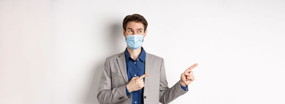Covid-19, pandemic and business concept. Excited young man in medical mask and suit looking left, pointing at logo with pleased face, white background.