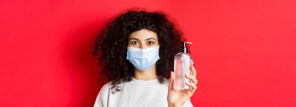 Covid-19, pandemic and quarantine concept. Young woman in medical mask showing bottle of hand sanitizer, demonstrate antiseptic, red background.
