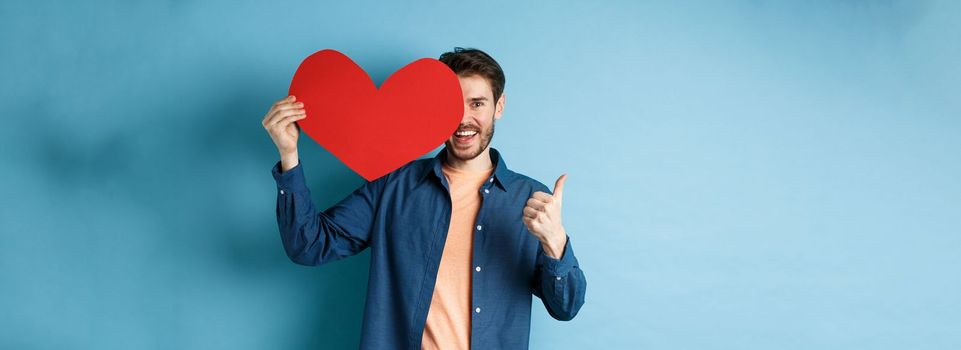 Happy man showing valentines heart and thumbs-up gesture, standing over blue background.