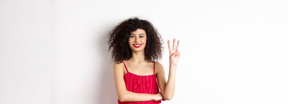 Beautiful lady with curly hair, wearing elegant red dress and showing number three, making order, standing over white background.