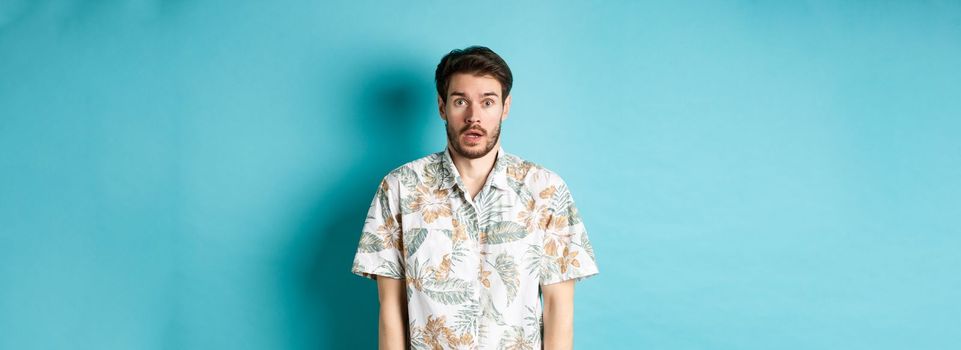 Image of shocked tourist guy drop jaw, gasping and looking startled, standing in hawaiian shirt on blue background. Copy space
