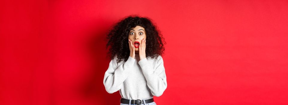 Portrait of shocked woman scream amazed, touching face and looking at camera at impressive promo offer, standing in sweatshirt on red background.