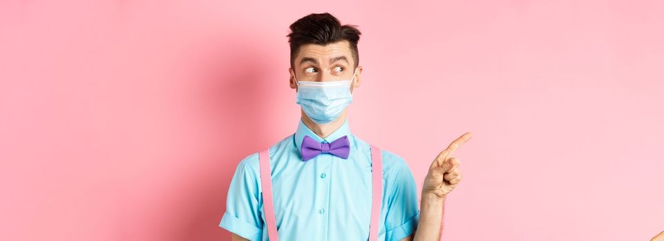 Covid-19, pandemic and health concept. Funny man in bow-tie and medical mask pointing finger left, looking aside curious, showing logo, standing on pink background.