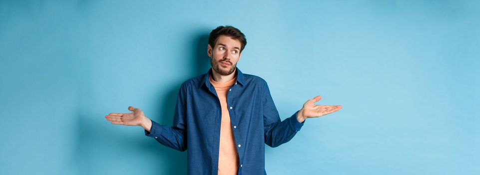 Indecisive young man with beard, look away and shrugging shoulders, know nothing, being clueless, standing on blue background.