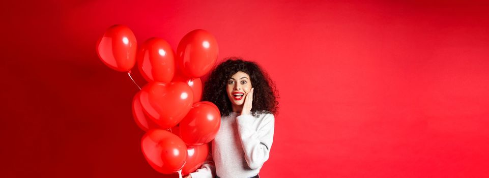 Valentines day. Surprised curly-haired girl holding heart balloons and looking amazed at camera, receive surprise gift on date from lover, standing on red background.