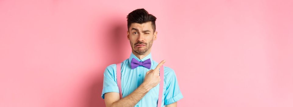 Childish young man acting sad and fake crying while pointing finger upper left corner, complaining on something, standing over pink background.