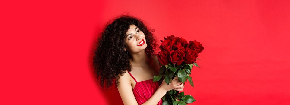 Happy beautiful woman in dress, holding flowers and smiling romantic, looking aside at logo, standing against red background.