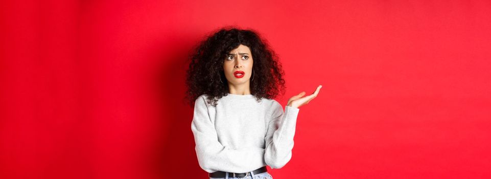 Annoyed and puzzled young woman with curly hair, raising hands up and looking aside, cant understand something strange, standing on red background.