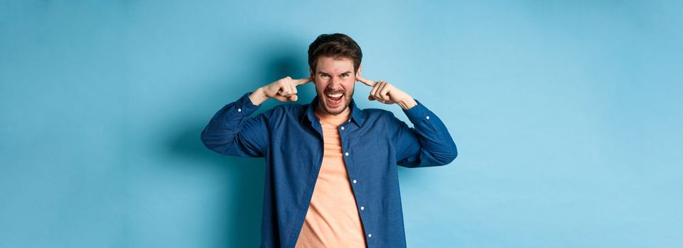 Angry young man shut ears to block annoying sound and shouting at camera, frowning and looking furious, standing on blue background.