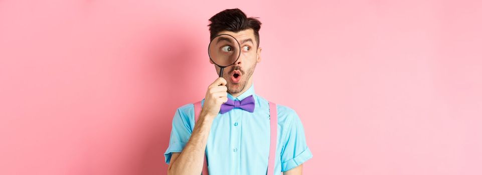 Surprised man saying wow and look aside through magnifying glass, checking out promo offer, standing over pink background. Copy space