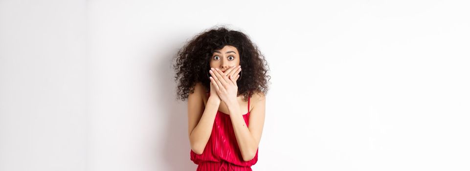Shocked woman covering mouth with hands and staring with disbelief at camera, witness something shocking, standing in red dress on white background.