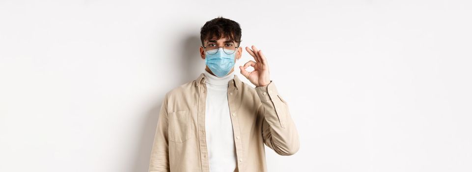 Covid-19, health and real people concept. Natural guy in glasses and medical mask show Ok sign, agree or approve something, standing on white background.