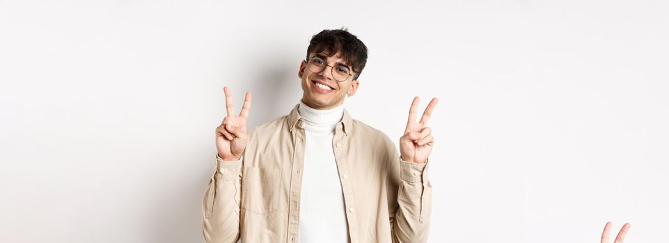 Handsome hipster guy in glasses, smiling with white teeth, showing peace or victory gesture, standing upbeat on white background.