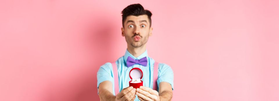 Valentines day. Funny young man pucker lips for kiss and showing engagement ring, making proposal, say marry me to lover, standing over pink background.