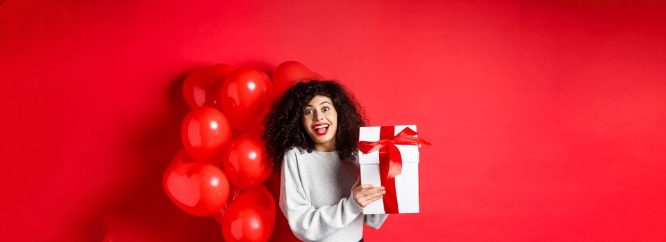 Surprised and happy woman holding valentines day gift from lover, standing near romantic hearts balloons and looking at camera amazed, red background.