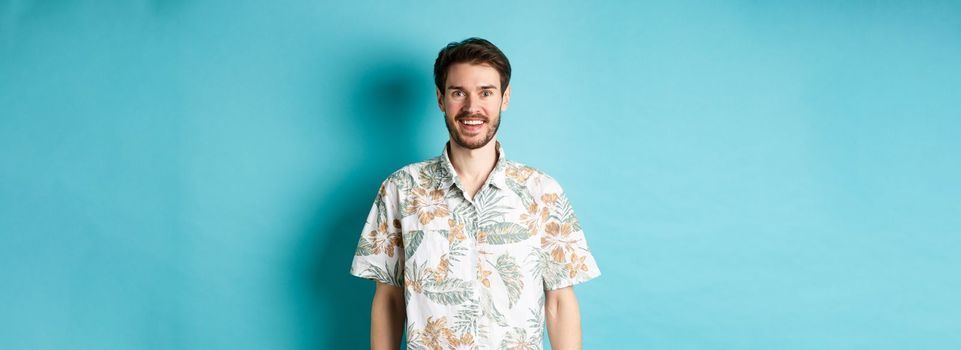 Cheerful caucasian guy going on vacation, wearing summer shirt and smiling, standing on blue background.