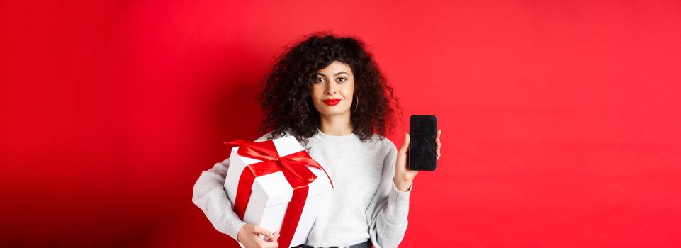 Beautiful woman with curly hair, showing shopping app on empty smartphone screen, holding gift wrapped in festive box, standing on red background.