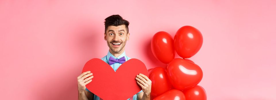 Valentines day concept. Cute young man in bow-tie showing big red heart postcard and say love you, smiling happy at camera, standing on romantic pink background.