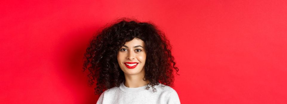 Close-up of beautiful lady with red lips and curly hair, smiling and looking happy at camera, studio background.