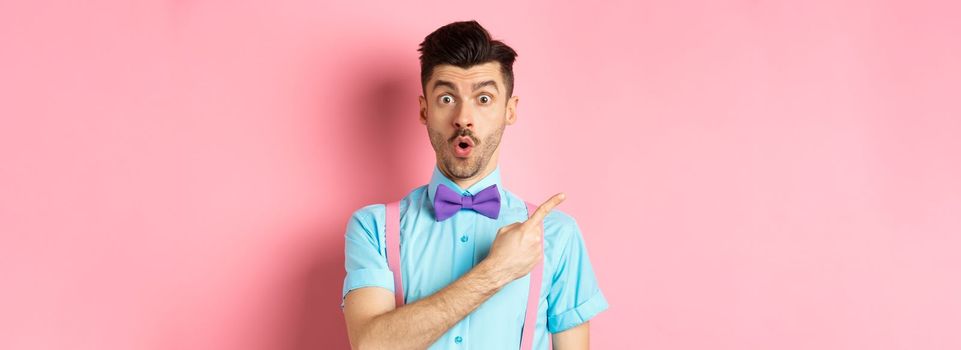 Check it out. Amazed caucasian man with moustache, wearing bow-tie, saying wow and pointing left, showing advertisement, standing on pink background.