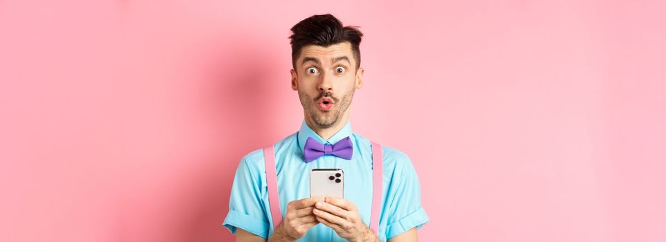 Online shopping. Intrigued guy in bow-tie, checking out internet promo offer on mobile phone, say wow at camera, holding smartphone, standing on pink background.