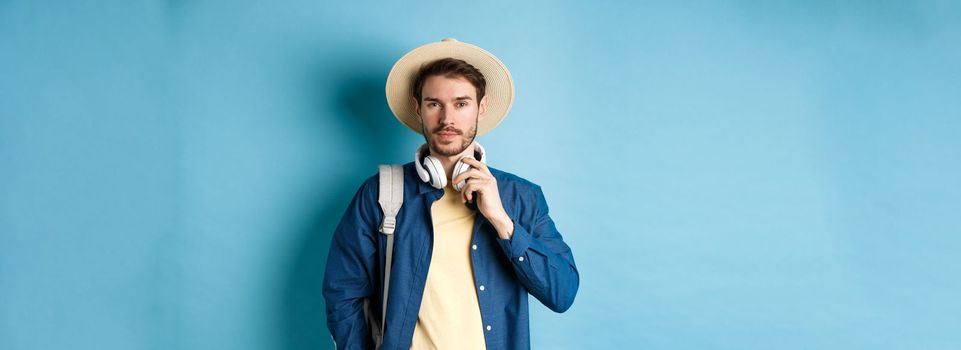 Handsome guy travelling with headphone, wearing straw hat and smiling, standing with backpack on blue background.