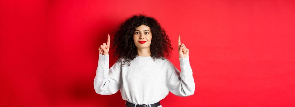 Image of cheerful smiling woman with curly hairstyle and red lips, pointing fingers up at empty space, showing advertisement, standing in casual clothes on studio background.