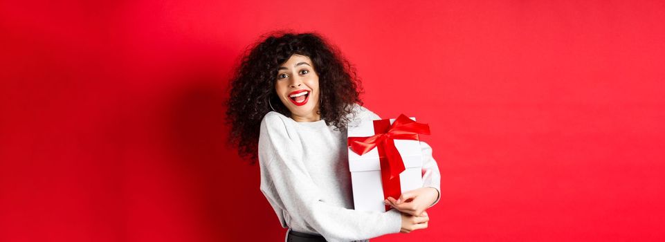 Valentines day. Happy girlfriend holding gift box from secret admirer, looking romantic at lover, receive present, standing on red background.