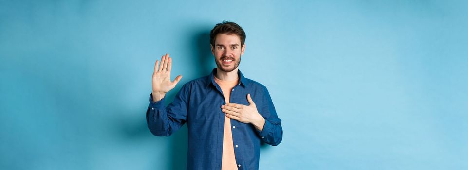 Honest young man smiling and making promise, holding hand on heart and arm raised, pledge or give oath, swearing to tell truth, standing on blue background.