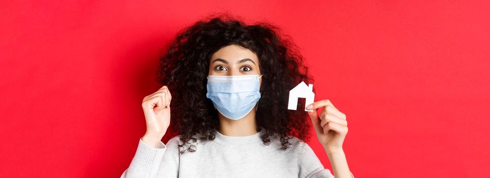 Real estate and covid-19 concept. Excited woman in medical mask showing small paper house cutout, standing on red background.