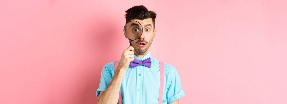 Funny guy look through magnifying glass with surprised face, seeing something interesting, standing on pink background.