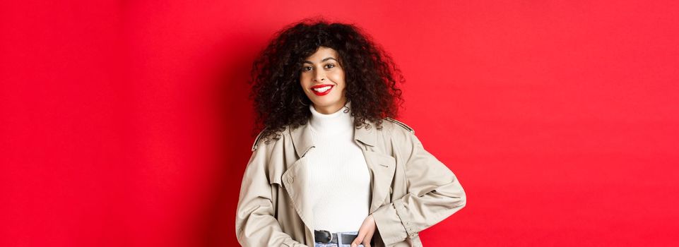 Modern woman with curly hair, wearing outdoor clothes, going for walk, standing against red background.