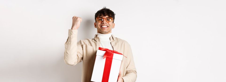 Cheerful guy saying yes as receiving gift, making fist pump and rejoicing, got present, standing on white background.