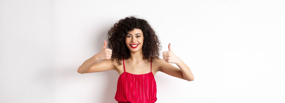 Attractive young woman recommending promo offer, showing thumb up and smiling, like product, standing in festive red dress on white background.