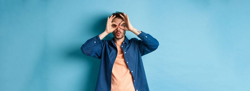 Funny young man looking through hand binoculars and say wow, checking out something interesting, standing amazed on blue background.