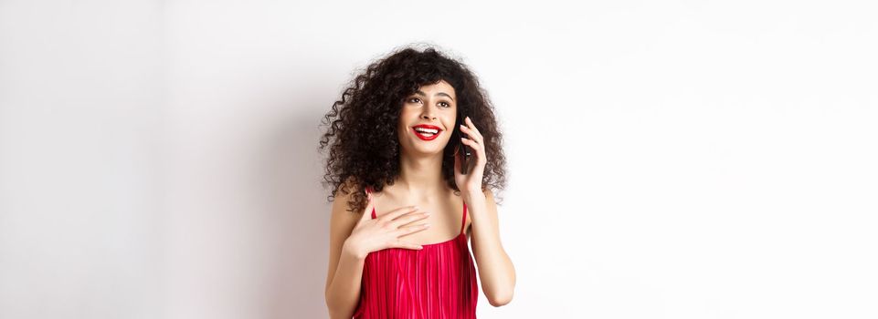 Beautiful lady with curly hair, wearing red dress and talking on phone, happy face, having pleasant conversation, standing on white background.