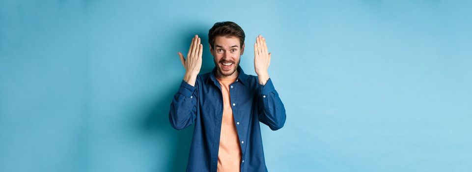 Happy young man open eyes and look surprised at gift, standing on blue background.