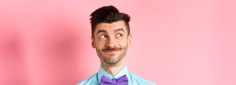 Close-up of handsome smiling guy with moustache, looking left at logo with pleased face, standing on pink background. Copy space