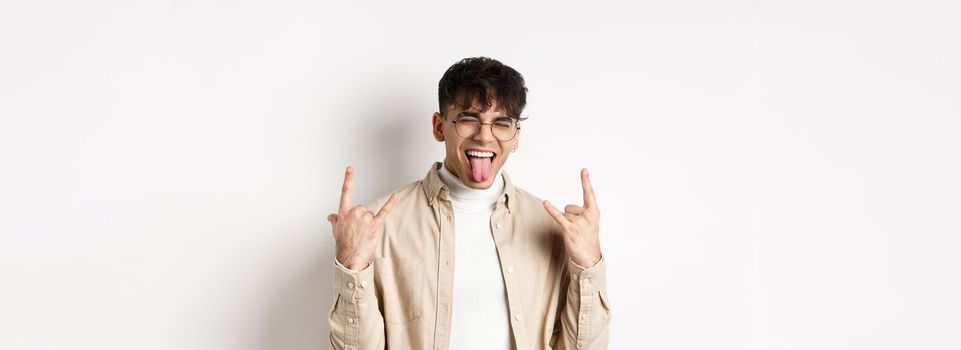 Happy handsome guy showing tongue and rock-on gesture, having fun and feeling upbeat, enjoying something good, standing on white background.