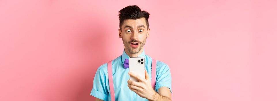 Impressed man reading mobile screen with amazed face, seeing online promo offer, standing over pink background. Copy space