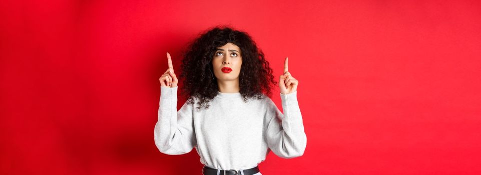 Concerned young woman with curly hair, frowning and looking doubtful, pointing up with hesitant or worried face, standing on red background.