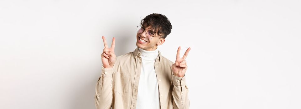 Real people. Handsome hipster guy in glasses showing peace gestures and smiling cute, standing on white background.