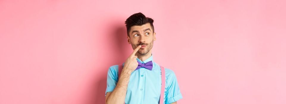 Image of handsome funny man in bow-tie looking thoughtful left and touching lip, thinking what to choose, making decision, standing over pink background.