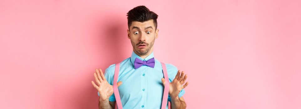Funny guy with moustache and bow-tie, adjusting his suspenders and looking down with confused and surprised face, standing over pink background.