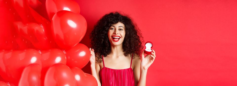 Happy beautiful lady feeling excited about marriage proposal, showing engagement ring and laughing, standing near heart balloons on red background.
